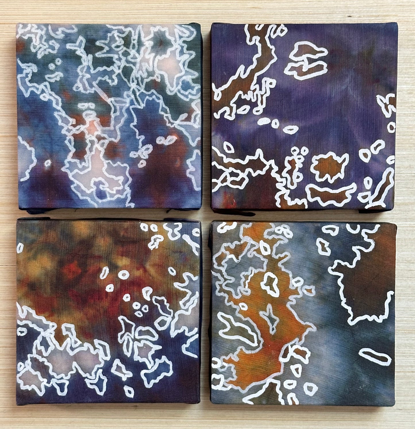 Four square silk paintings with white abstract design on a wooden surface