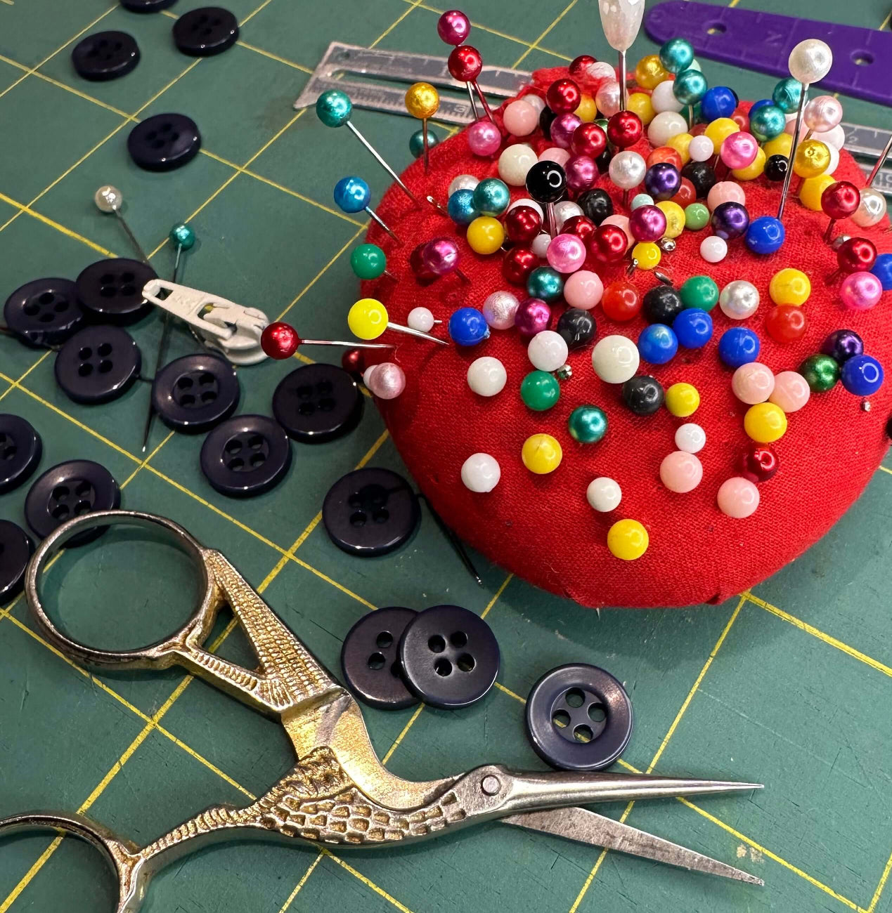 Various sewing buttons and fabric patches are displayed on a table with a pair of scissors and a red sewing project in the background.