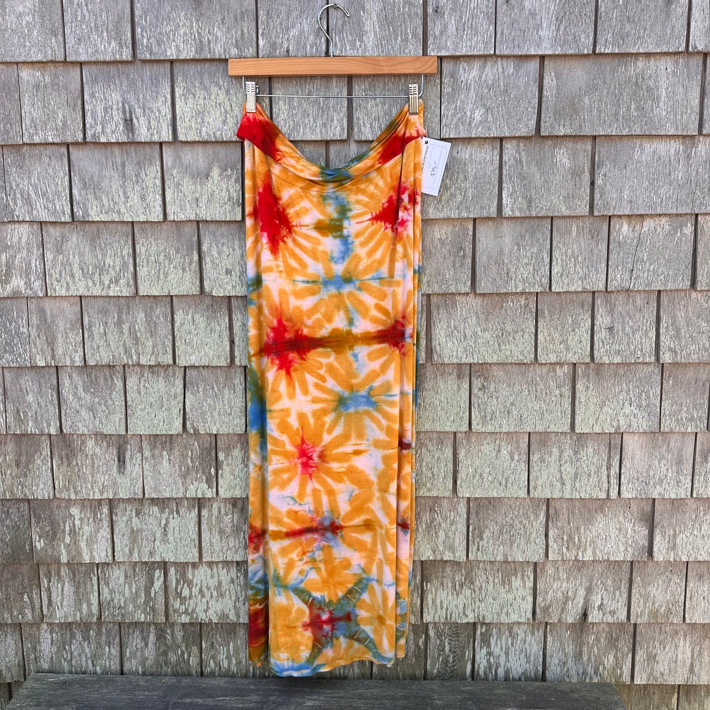 A maxi skirt with a vibrant tie-dye starburst pattern in orange, red, blue, and green, hanging from a wooden hanger against a gray shingled wall.