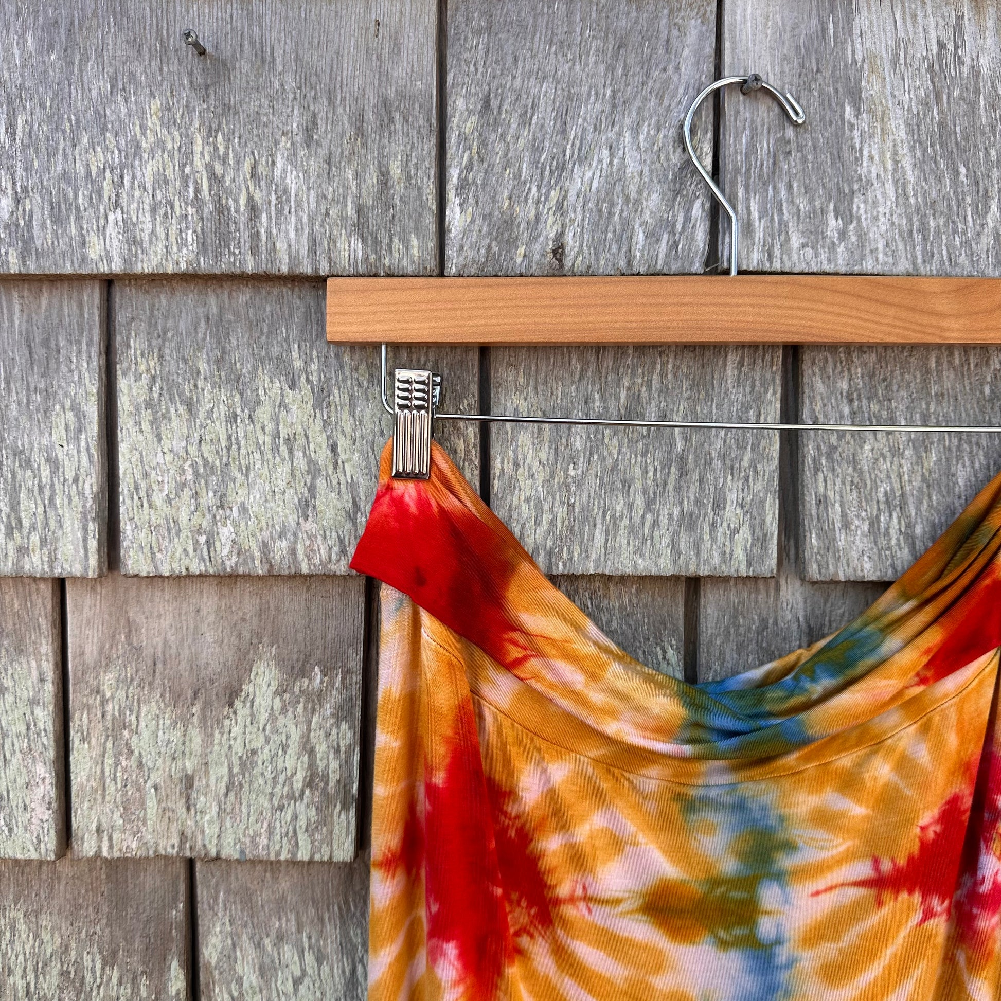 A colorful tie-dye skirt with red, orange, yellow, green, and blue patterns hanging from a wooden hanger against a weathered gray wooden shingle wall.