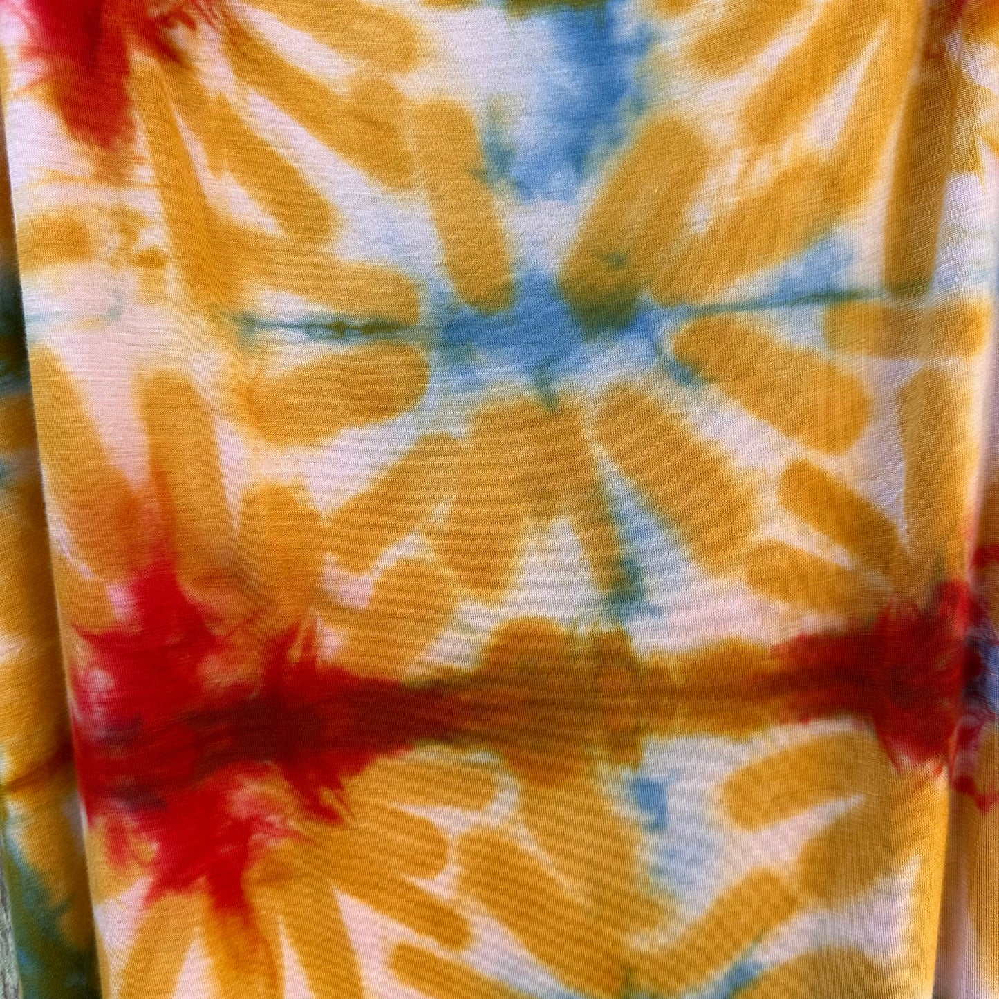 A close-up of a tie-dye fabric with a radial burst pattern in mustard yellow, light blue, and red on a white base.