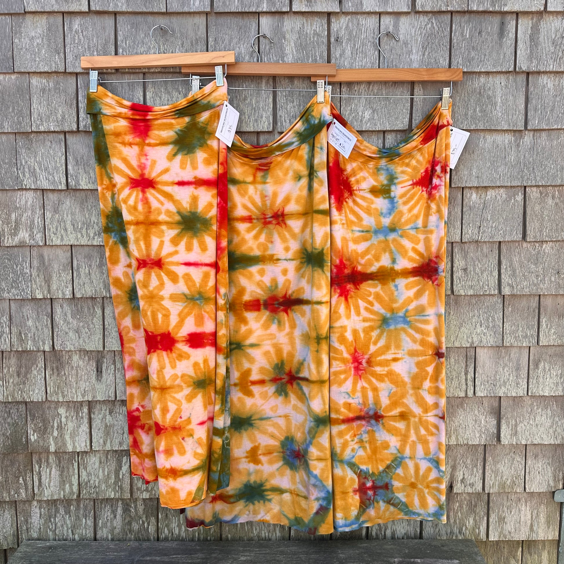 Three tie-dye maxi skirts  in orange, yellow, green, and red hang on wooden hangers against a gray wood shingle wall.