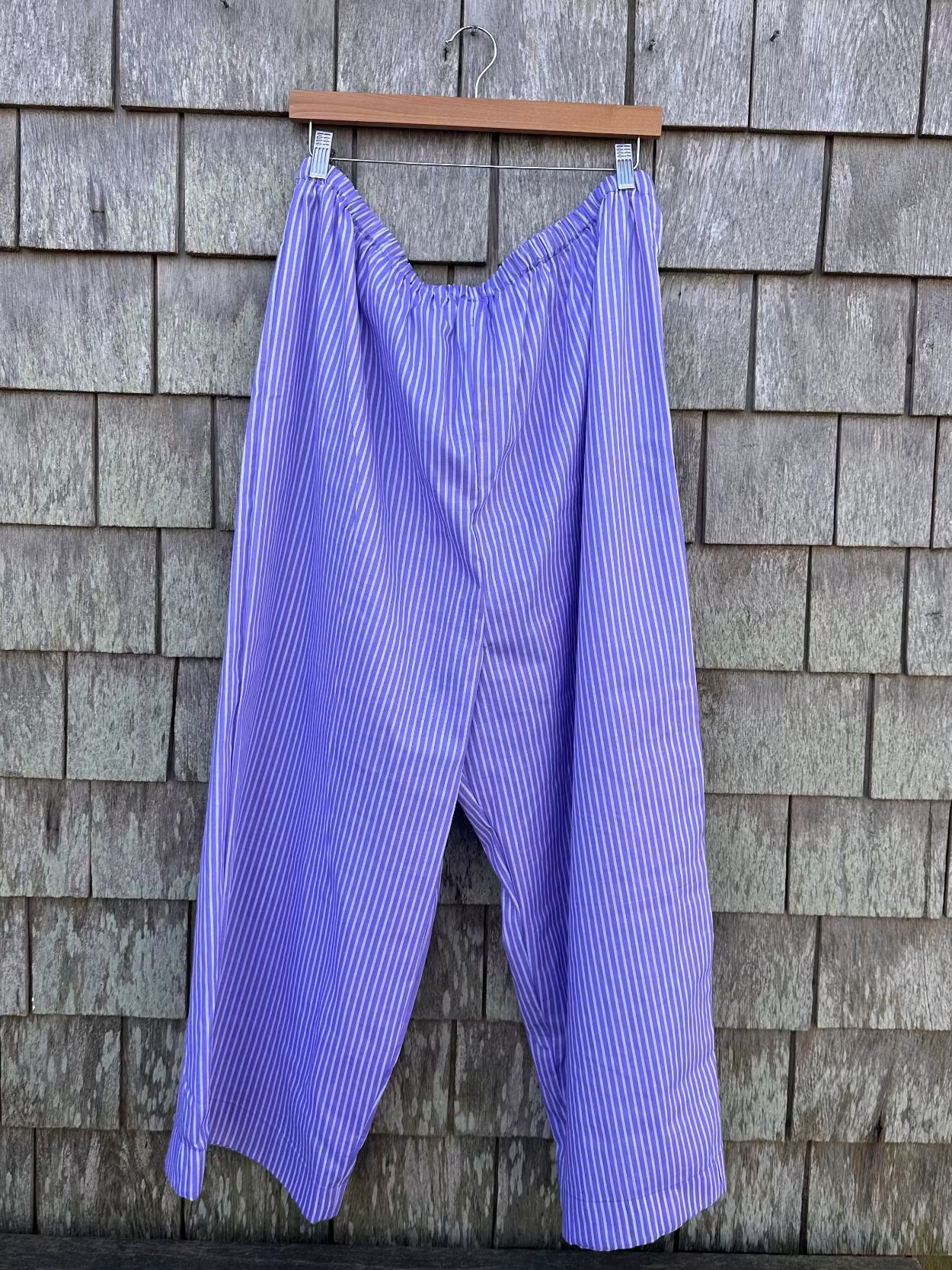 Purple striped pants hanging on a wooden hanger against a gray shingled wall.